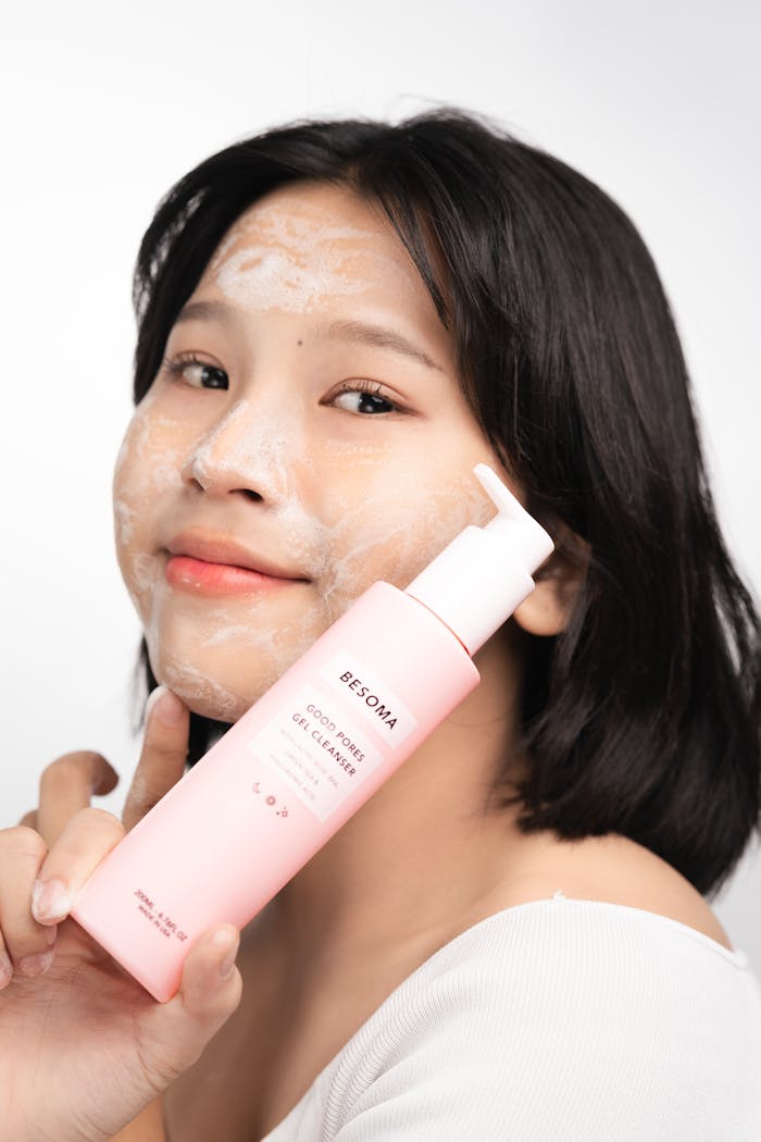 Smiling woman with cleanser foam showcases skincare bottle in studio setting.